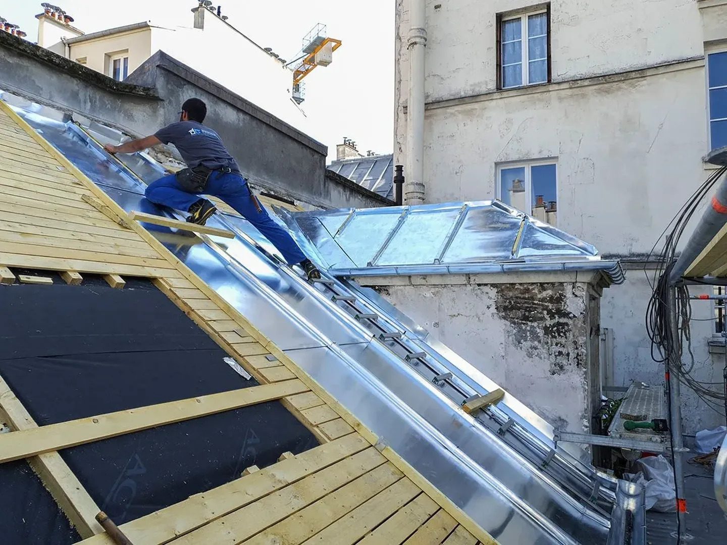 Un ouvrier installe des panneaux métalliques à côté de puits de lumière sur un toit en pente. à Paris — Couvreur — par ADH75 Couverture 🆘 Urgence Fuite Toiture - couvreur IDF - Service Dépannage - Couvreur Paris entreprise de couverture à Paris